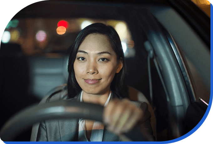 Asian woman smiling while driving a car at night, illuminated by ambient street lights.