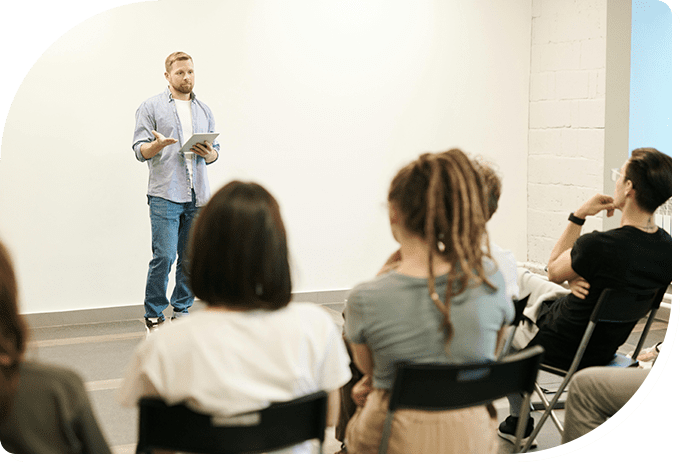 A man standing and speaking to a seated group in a bright room, holding a tablet, with attentive listeners facing him.